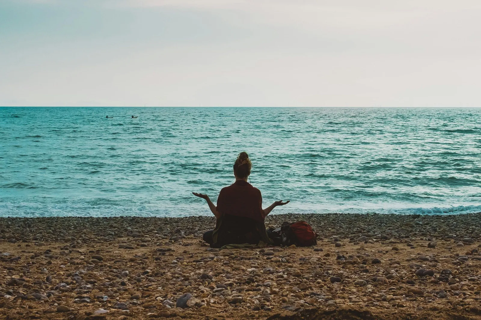 Person sitzt in Meditation auf einem Kiesstrand und blickt aufs offene Meer, ein Rucksack neben ihr, bewölkter Himmel.