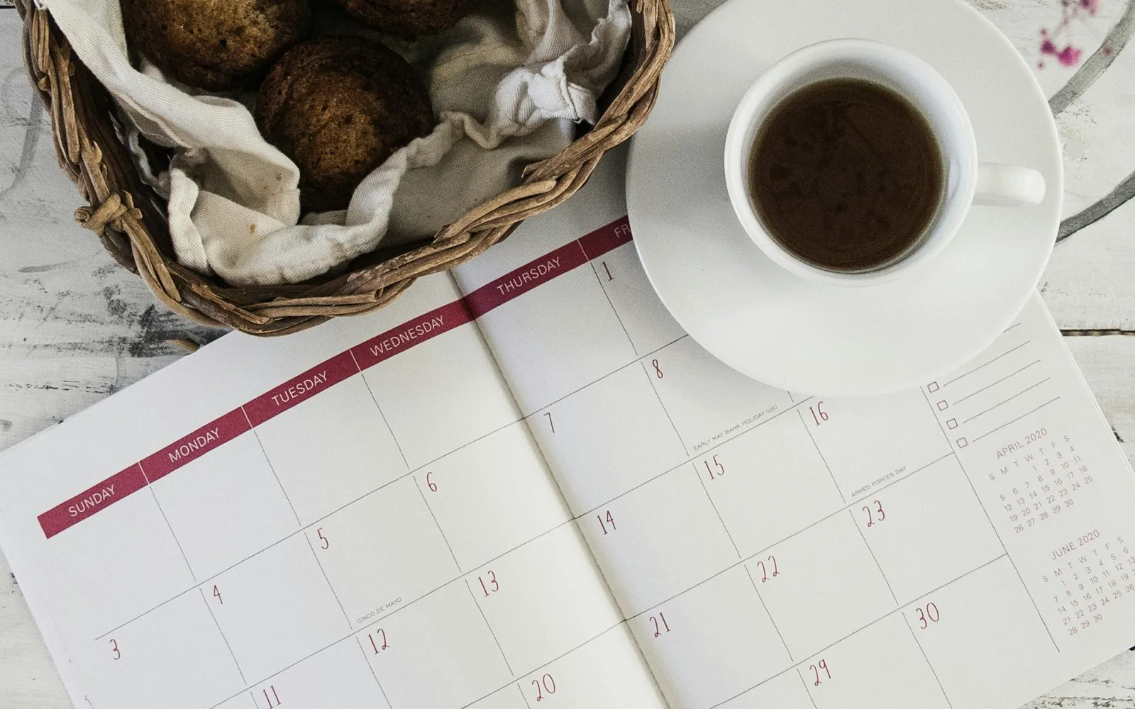 Flach liegender Papier-Monatskalender neben einer weißen Kaffeetasse und einem Korb mit Gebäck auf einer rustikalen Holzoberfläche.