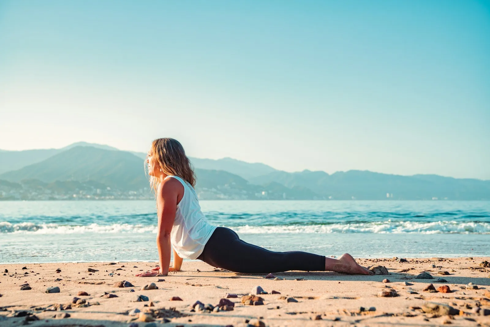 Frau in Upward-Dog-Yoga-Pose auf einem Sandstrand mit Bergen und Ozean im Hintergrund unter einem klaren Morgenhimmel.