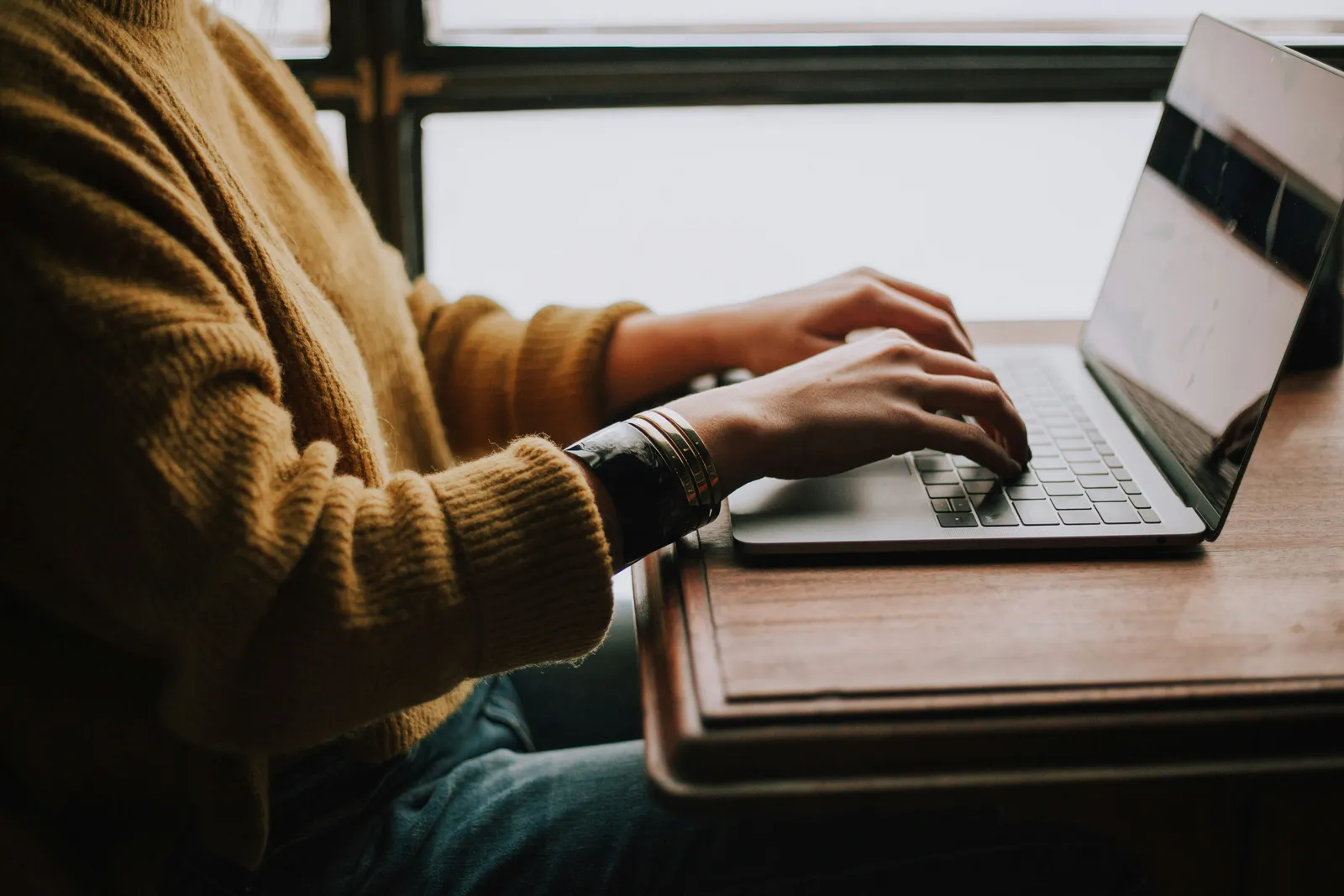 Frau im senfgelben Strickpullover tippt an einem Laptop auf einem Holztisch vor einem hellen Fenster, das Gesicht ist nicht im Bild.
