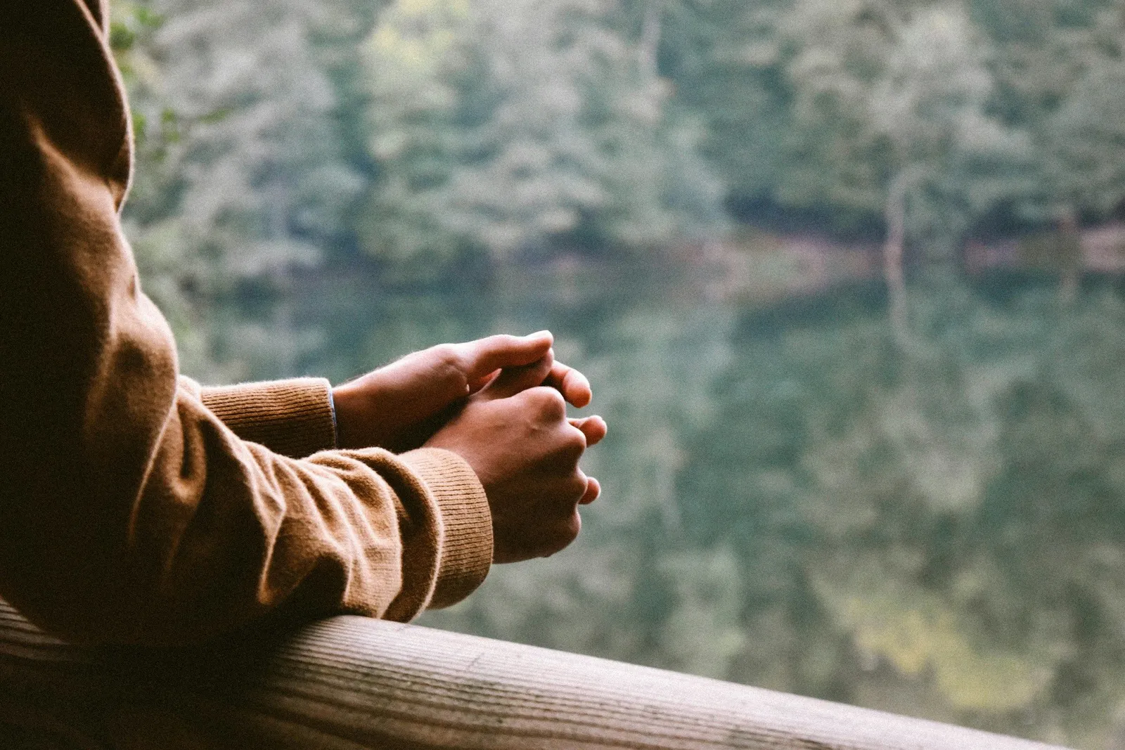 Person in braunem Pullover steht an einem Holzgeländer mit gefalteten Händen und blickt ruhig auf einen stillen Waldsee in der Ferne.