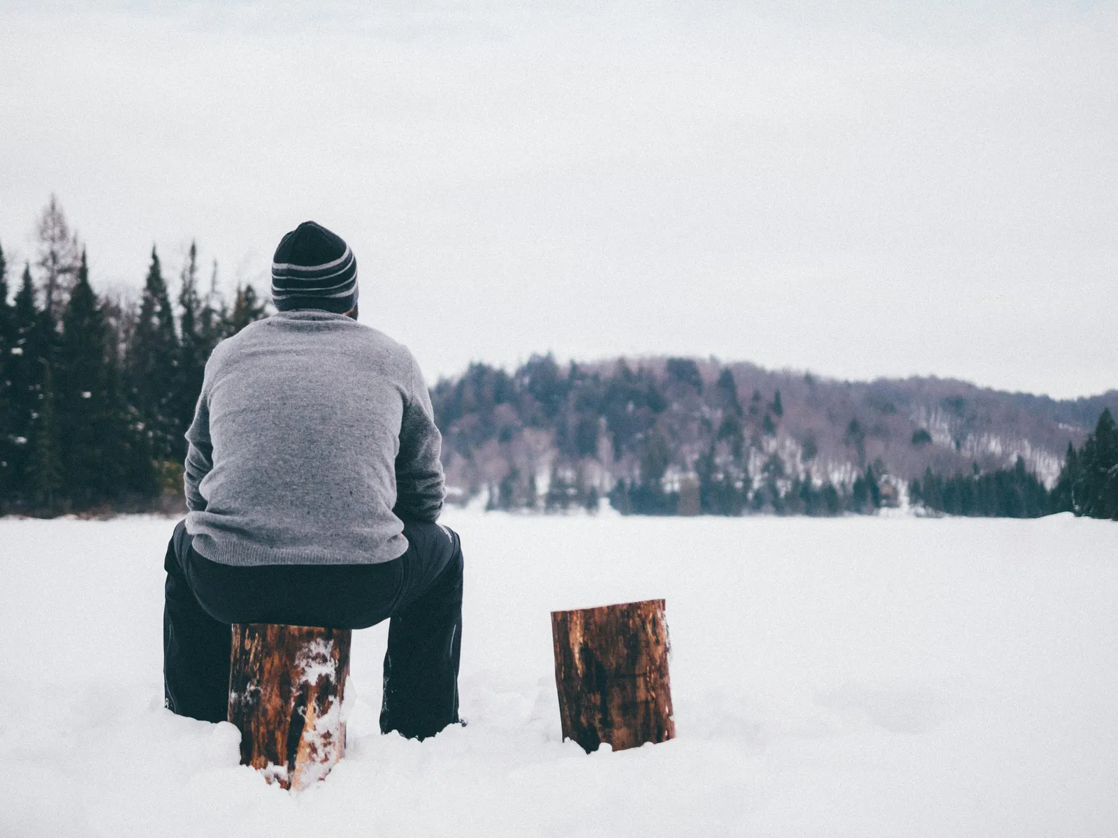 Person in Beanie und grauem Pullover sitzt allein auf einem zugefrorenen See, Blick auf einen Winterwald.