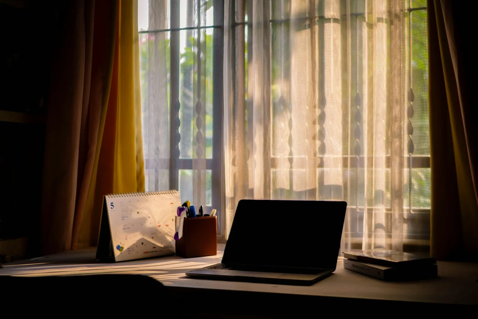 Ein Laptop steht aufgeklappt auf einem sonnenbeschienenen Holzschreibtisch am Fenster, daneben ein Kalender und eine Ordnungsbox, im Hintergrund ein grüner Garten.