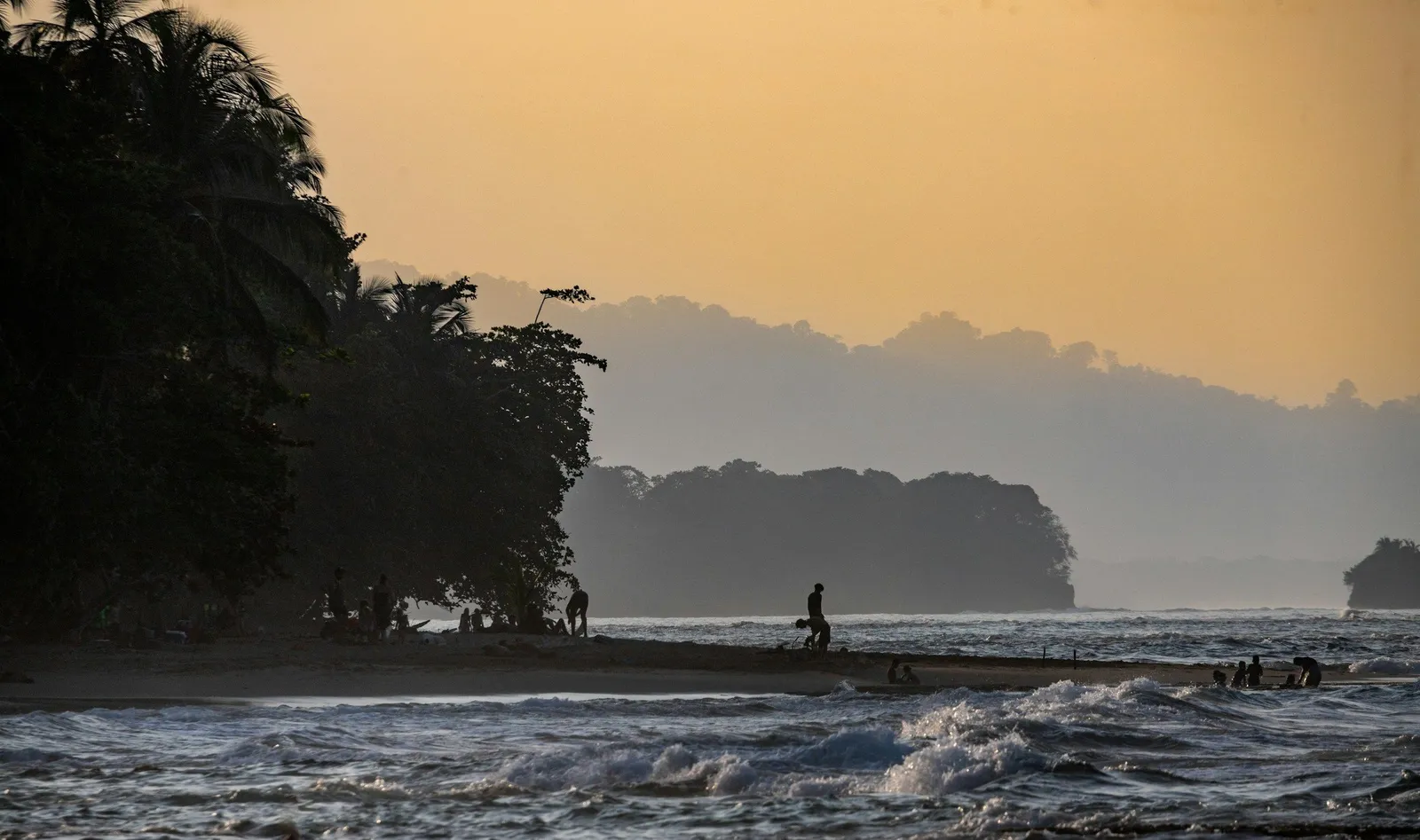 Tropischer Strand bei goldenem Abendlicht mit Silhouetten-Palmen und Menschen im Wasser an der Pazifikküste Costa Ricas.
