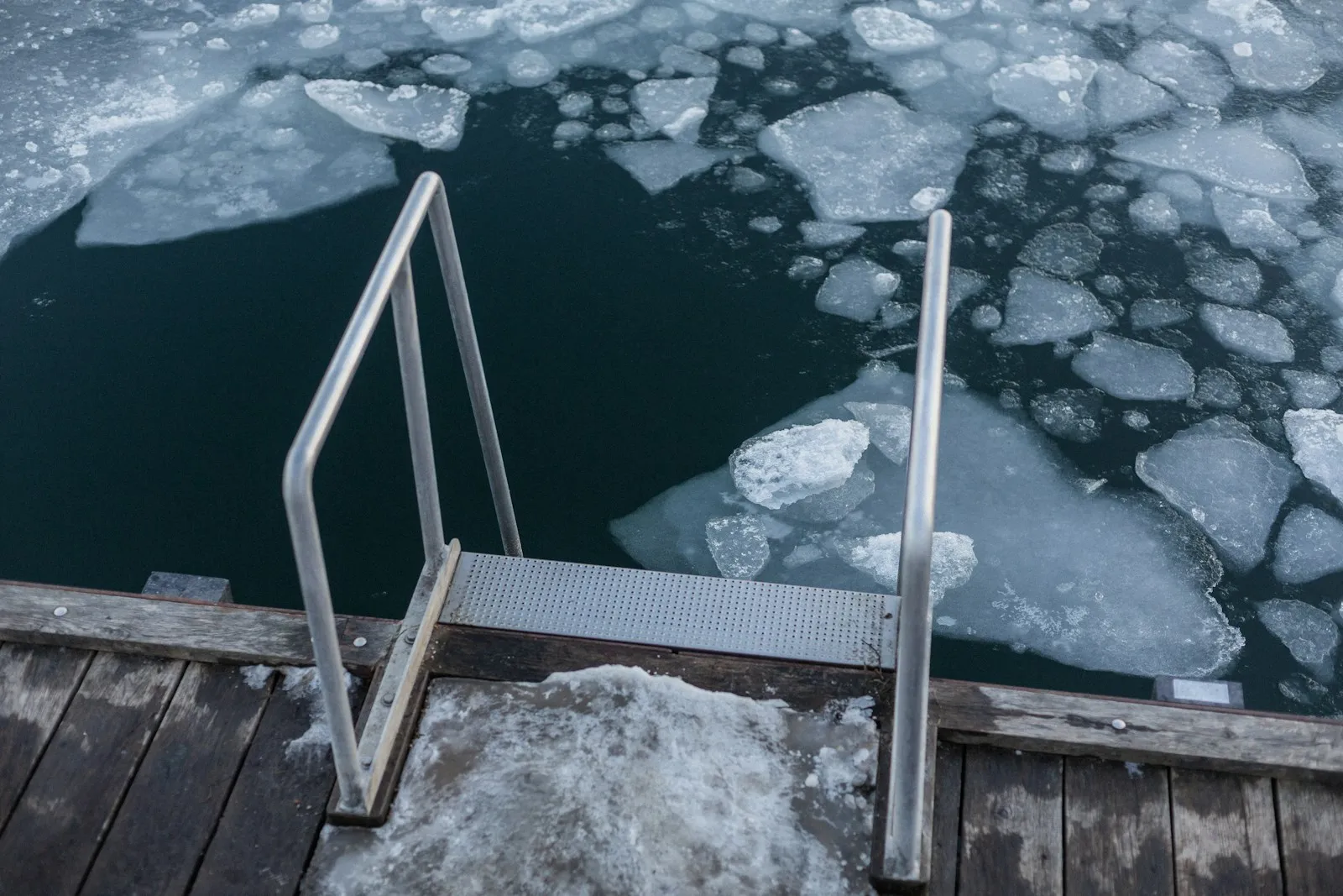 Eine Edelstahlleiter mit gelochter Metallstufe führt von einem verschneiten Holzsteg hinab in dunkles Wasser, auf dem zerbrochene Eisschollen treiben.