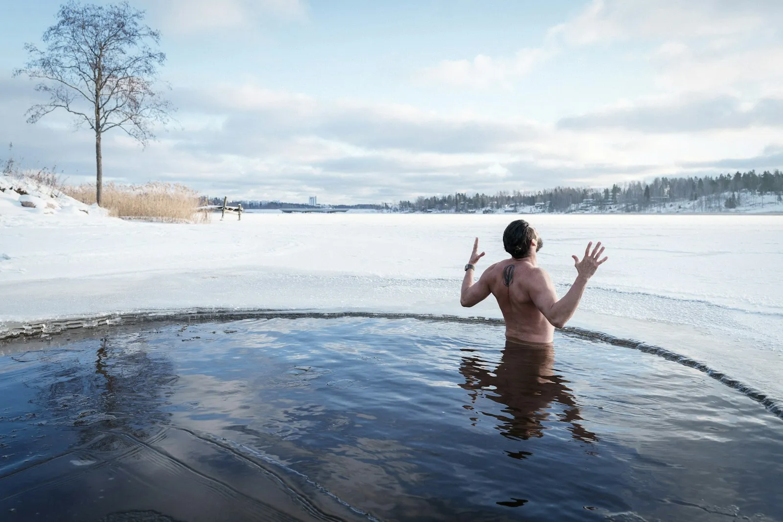 Ein Mann mit nassem Haar und Tattoo am Oberarm steht bis zur Hüfte in einem runden Eisloch in einem zugefrorenen, verschneiten See und hebt die Hände in die Luft, links ein kahler Baum am Ufer, im Hintergrund ein Waldstreifen unter hellem Winterhimmel.