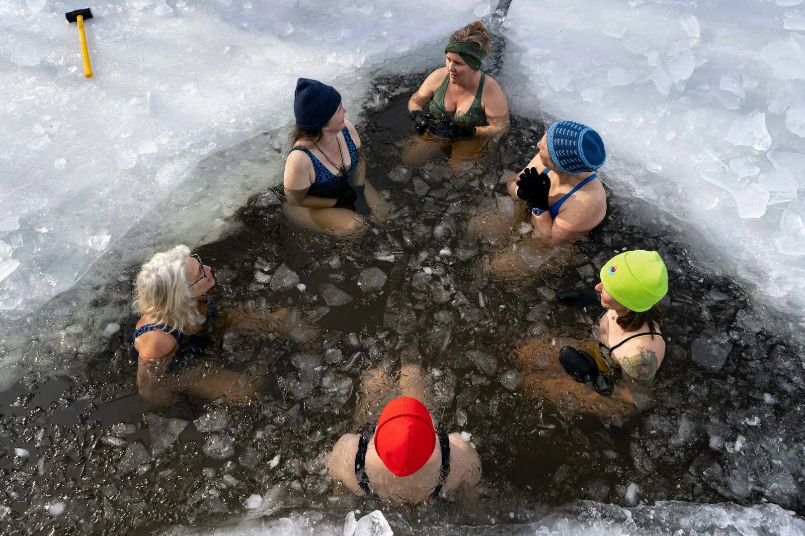 Sechs Frauen mit bunten Mützen und Neoprenhandschuhen sitzen im Kreis in einem Eisloch voller Eisbrocken, von oben fotografiert, am Rand liegt ein Vorschlaghammer auf dem Eis.