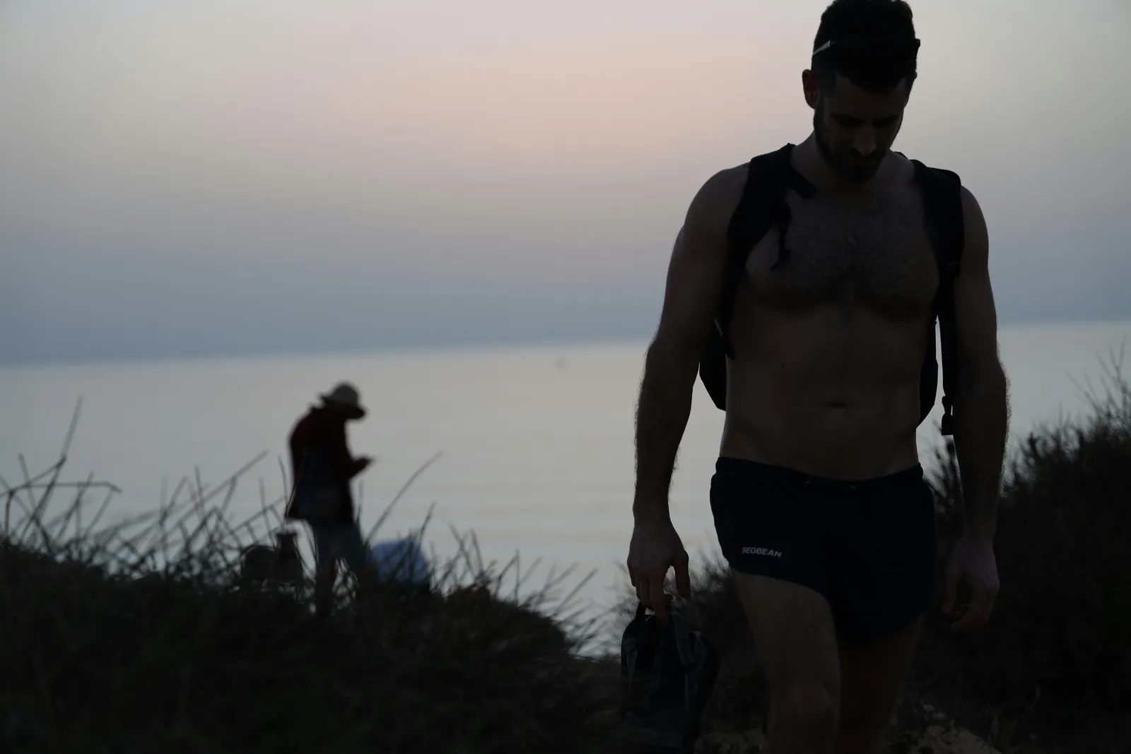 Hemdloser Mann mit Rucksack geht einen Küstenpfad hinauf in der Dämmerung, eine zweite Figur im Abstand hinter ihm