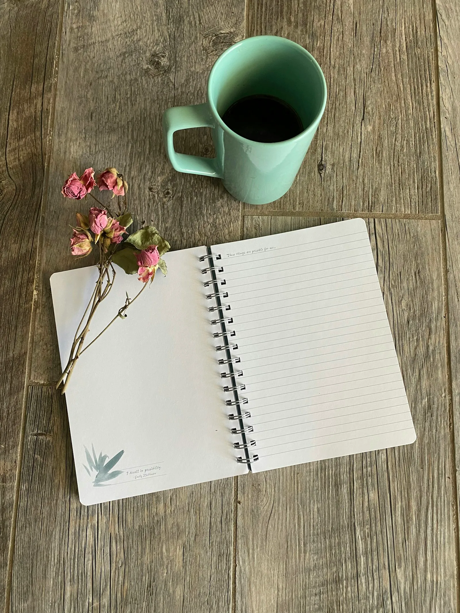 Ein aufgeschlagenes Spiralnotizbuch mit linierten Seiten liegt auf einem grauen Holztisch, daneben ein Zweig getrockneter Rosen. Oben steht ein mintgrüner Becher mit einem dunklen Getränk.