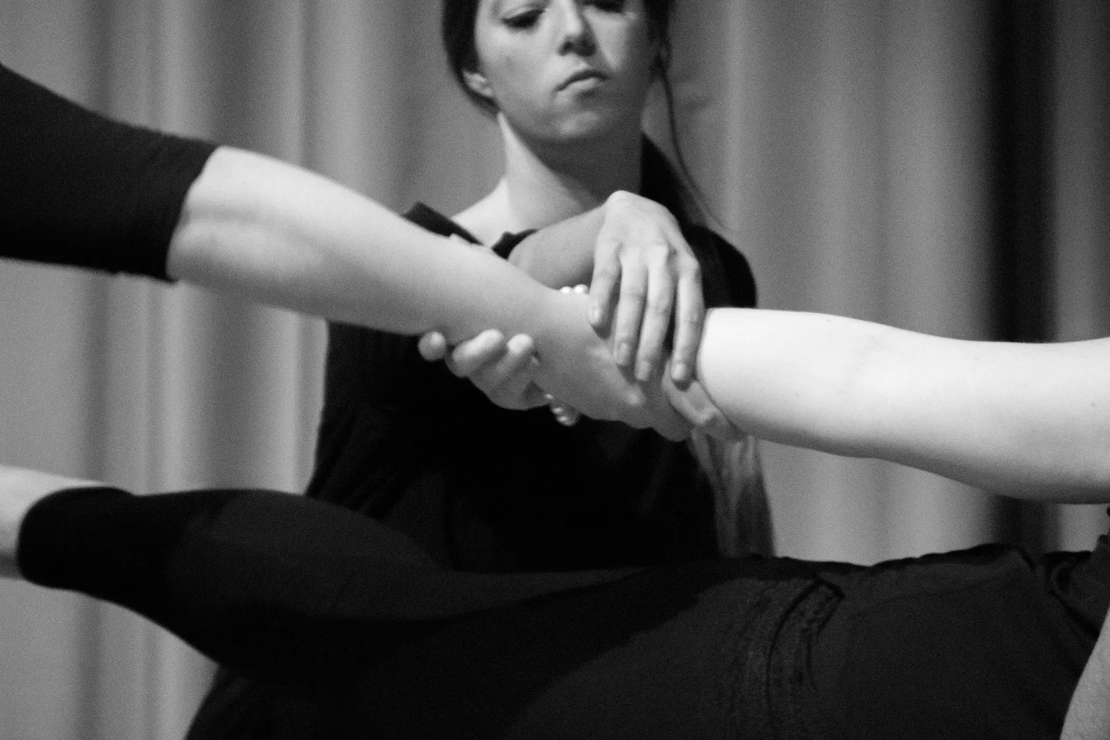 Schwarz-Weiß-Aufnahme einer Frau, die bei einem Bewegungsworkshop das ausgestreckte Bein einer anderen Person stützt. Konzentriert, körpernah, instruktiv.