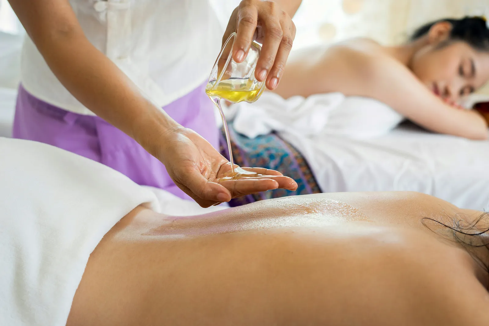 Massagetherapeutin gießt gelbes Öl in ihre Handfläche über den Rücken einer Klientin bei einer Spa-Behandlung, im Hintergrund eine weitere Frau auf einer Liege.