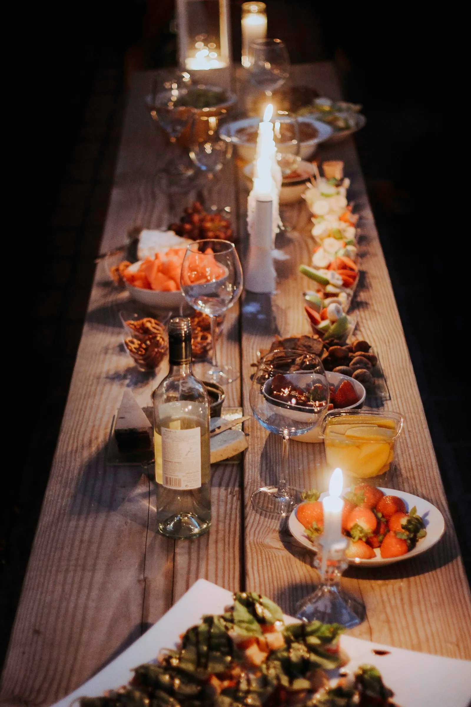 Eine lange Holztafel mit Kerzen, Weinflaschen und frisch zubereiteten Gerichten, beleuchtet in warmem Abendlicht.
