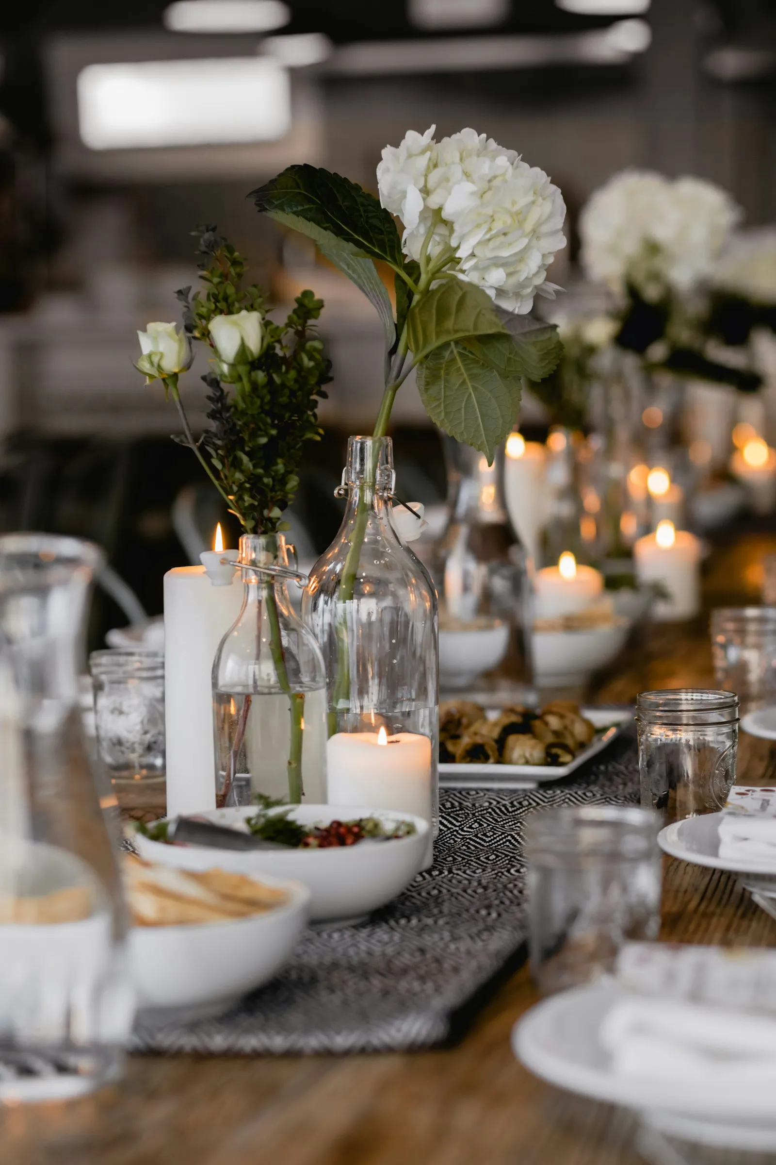 Eine Nahaufnahme eines eleganten Tischgedecks mit weißer Kerze im Glas, Blumen und unscharfem Kerzenlicht im Hintergrund.