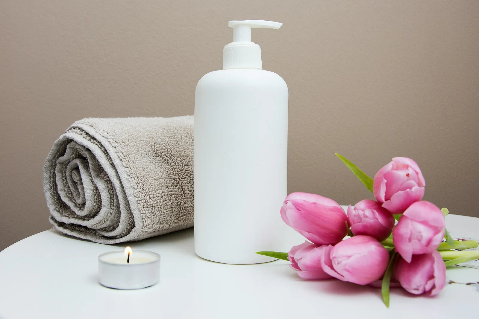 Ein Handtuch, eine Spa-Pumpflasche und rosa Tulpen liegen in ruhigem Licht auf einer hellen Fläche, dazu eine brennende Kerze.