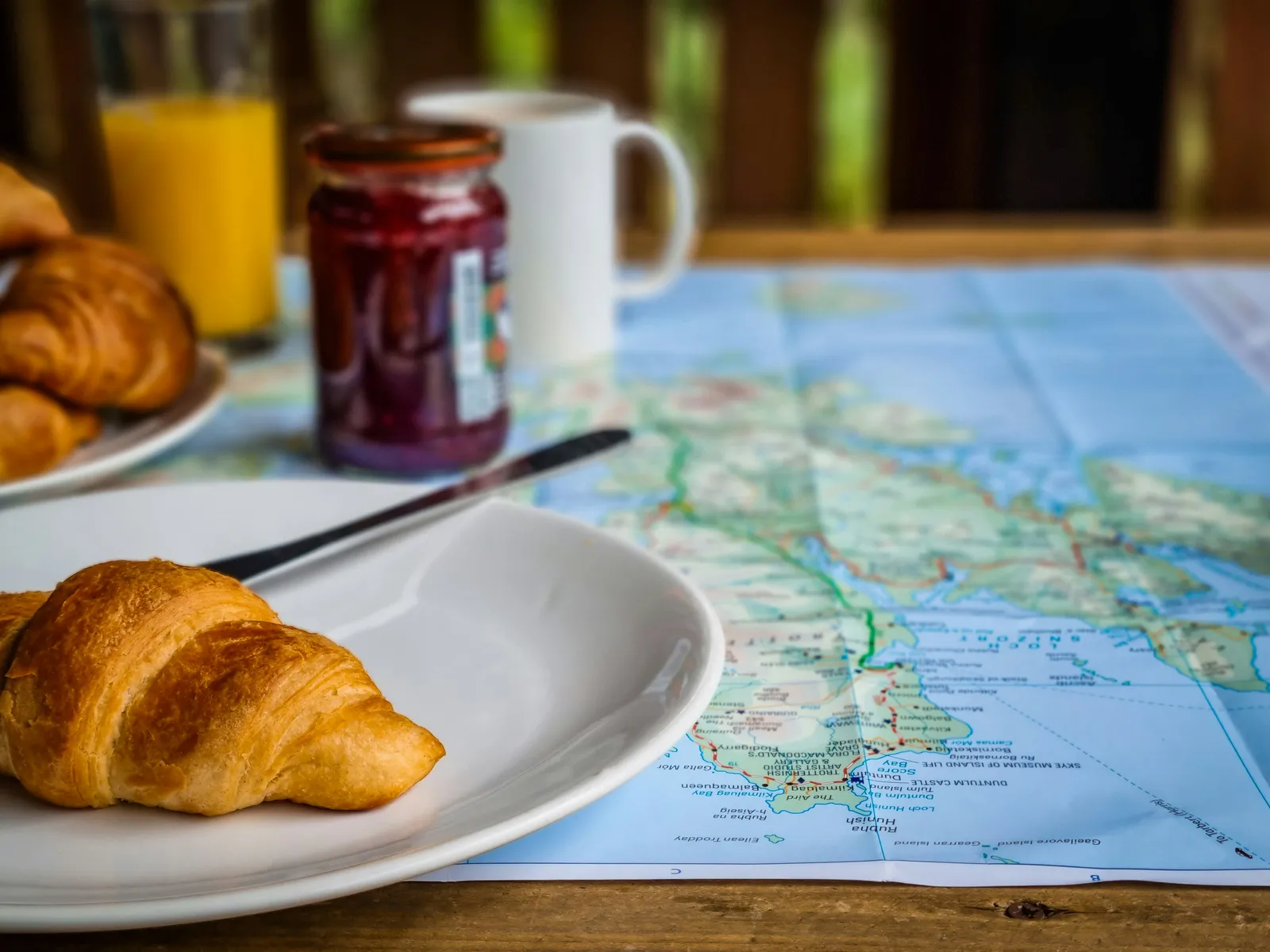 Frühstücksszene mit Croissant, Marmeladenglas, Kaffeetasse und Orangensaft auf einem Holztisch, daneben eine aufgeschlagene Straßenkarte.