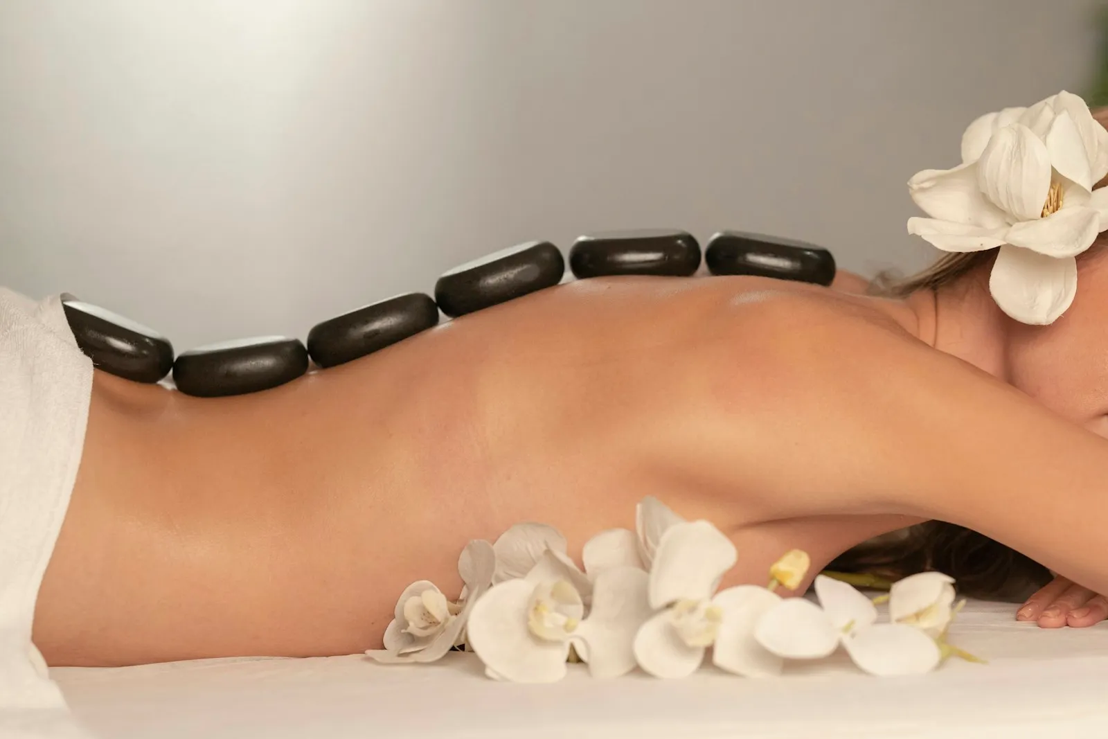 Glatte schwarze Steine liegen in einer Reihe entlang der Wirbelsäule einer Frau auf einer Massageliege, vorne weiße Orchideenblüten.