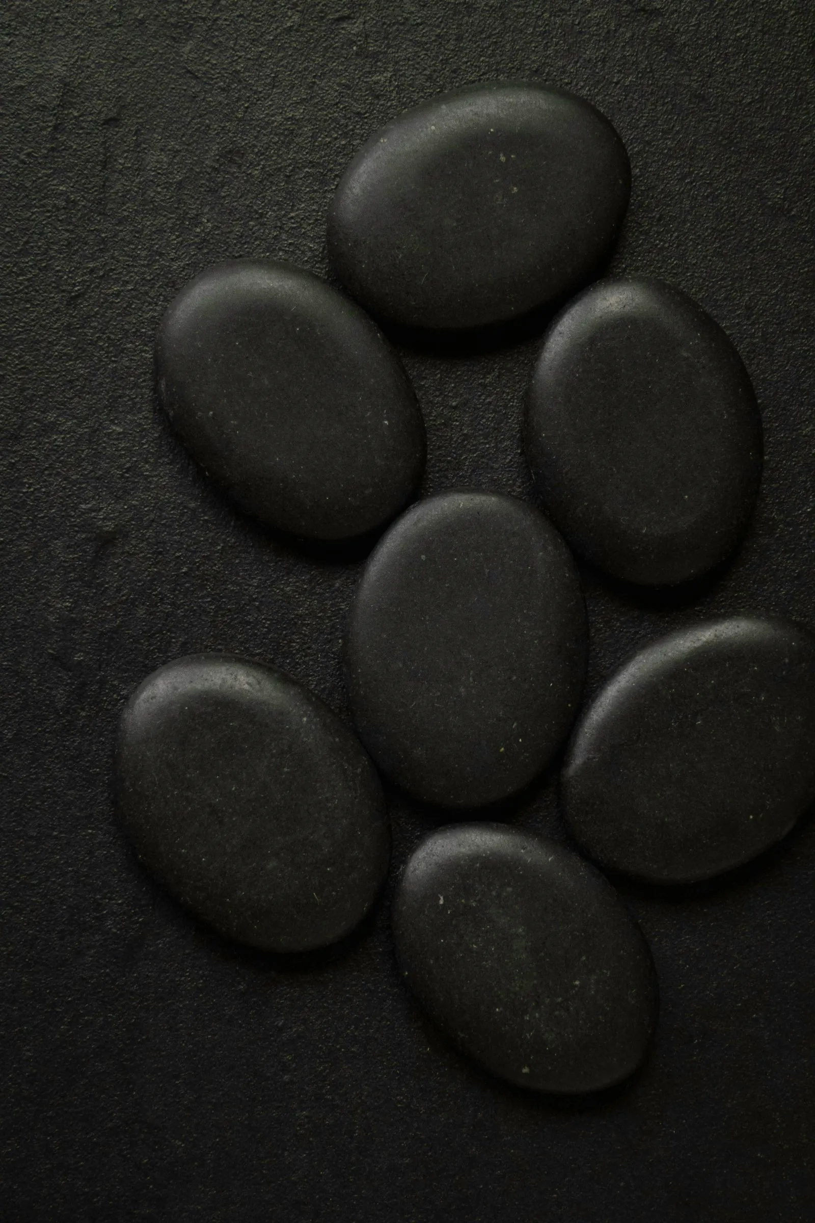 Sieben flache, glatte schwarze Steine liegen dicht nebeneinander auf dunklem Untergrund.