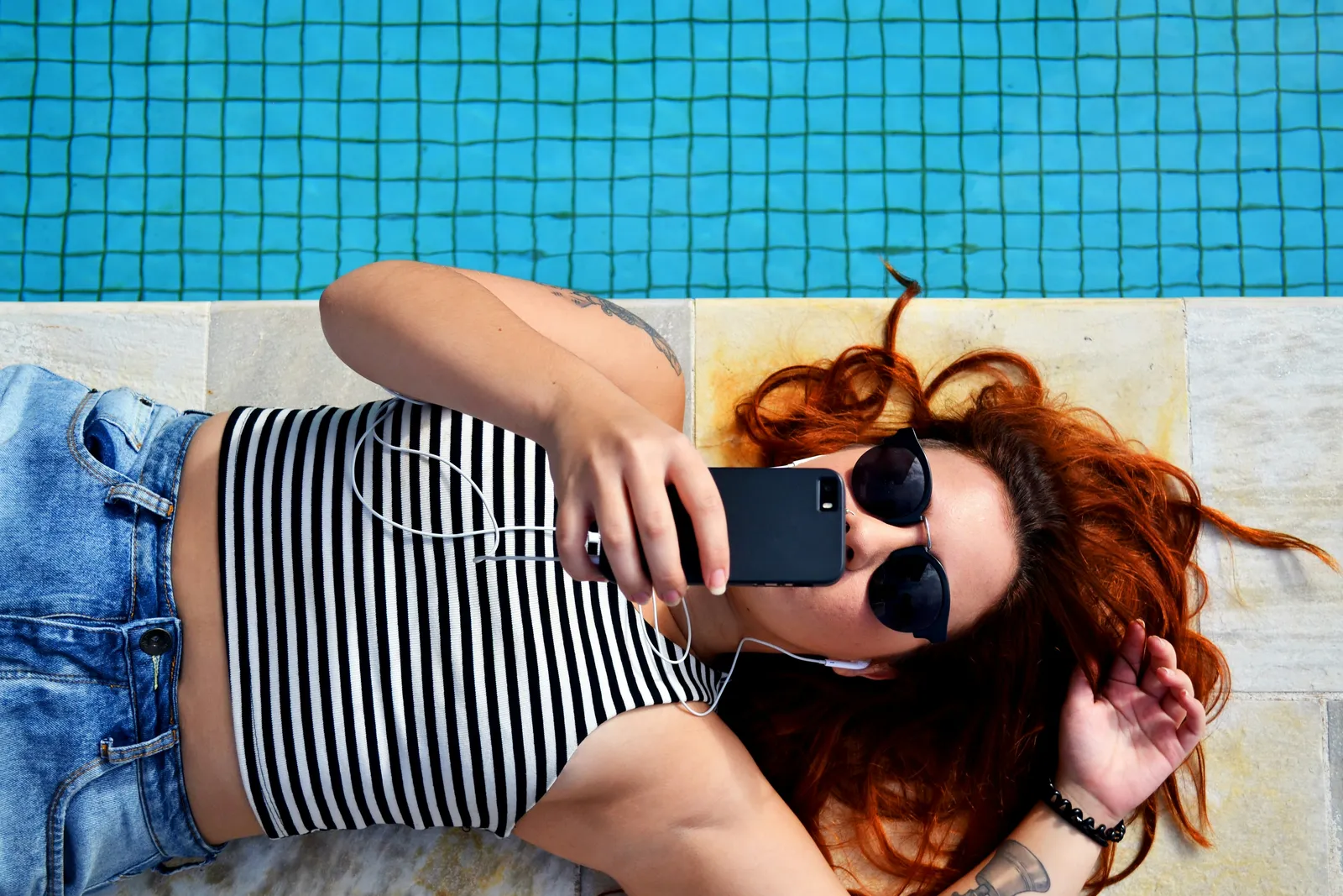 Blick von oben auf eine rothaarige Frau, die in der Sonne auf einem Handtuch am Pool liegt und ein Selfie mit dem Handy macht, Sonnenbrille auf.
