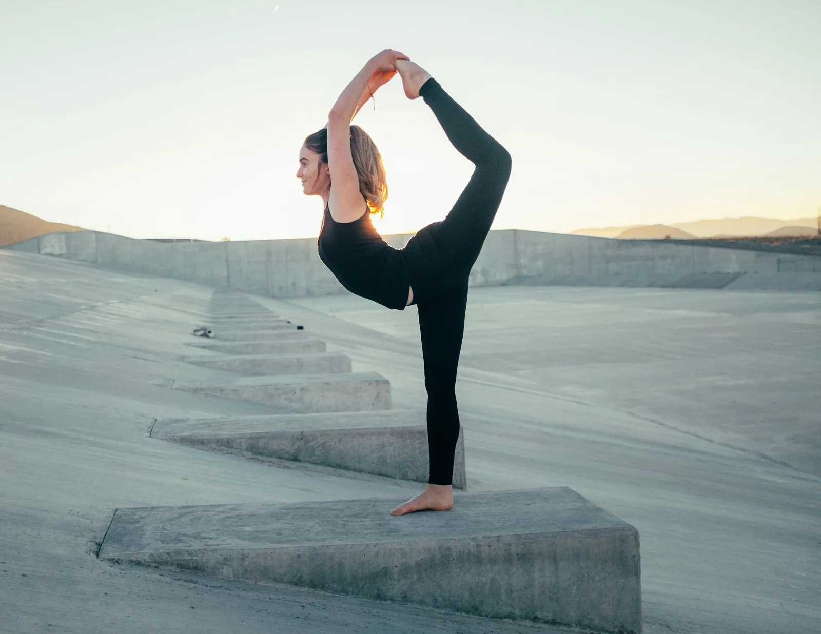 Frau in Stehbogen-Yogapose balanciert auf einem Bein auf einer Betonstufe in der Dämmerung, verschwommener Stadthintergrund