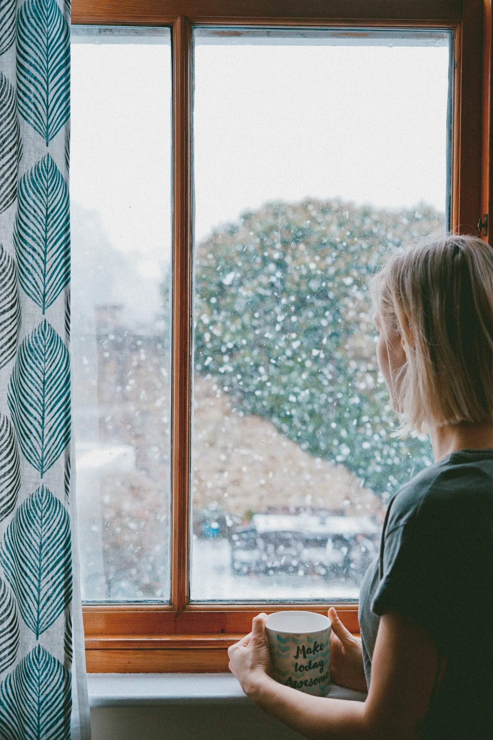 Eine Frau sitzt an einem großen, regenverhangenen Fenster, eine Tasse in der Hand, und blickt nachdenklich auf einen nebligen, bewaldeten Hügel.