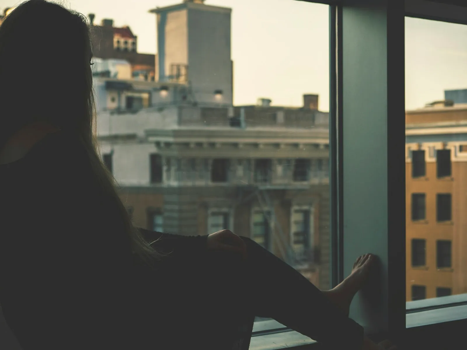 Eine Frau sitzt an einem großen Fenster im goldenen Abendlicht, silhouettiert vor dem Schein, der Blick auf eine ferne Stadtsilhouette in warmen Abendtönen gerichtet.