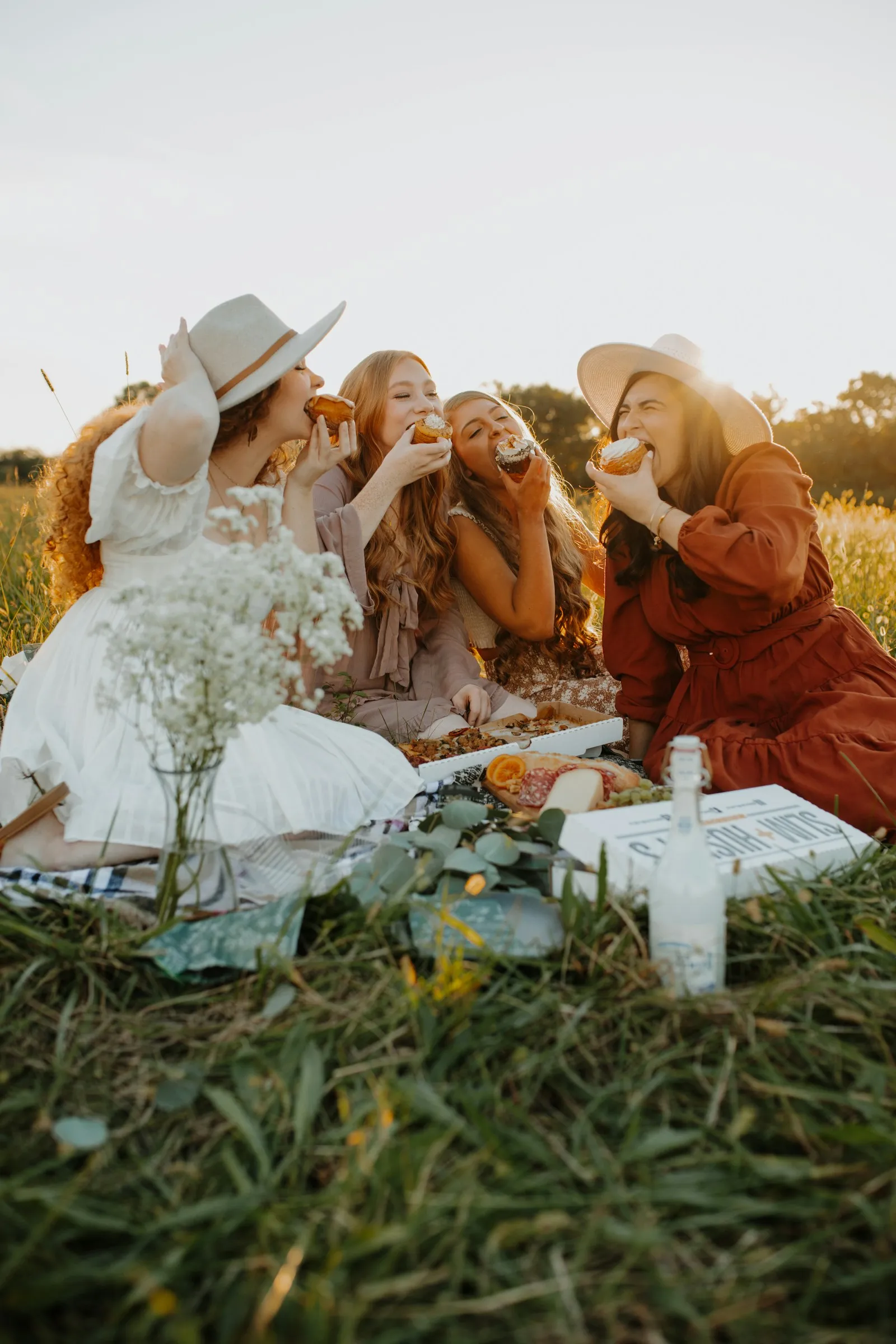 Vier Frauen in langen Kleidern, zwei davon mit Hüten, sitzen im Abendlicht auf einer Wiese um eine Picknickdecke und beißen lachend in Donuts. Vorn ein Glas mit Schleierkraut, eine Glasflasche und Pizzakartons.