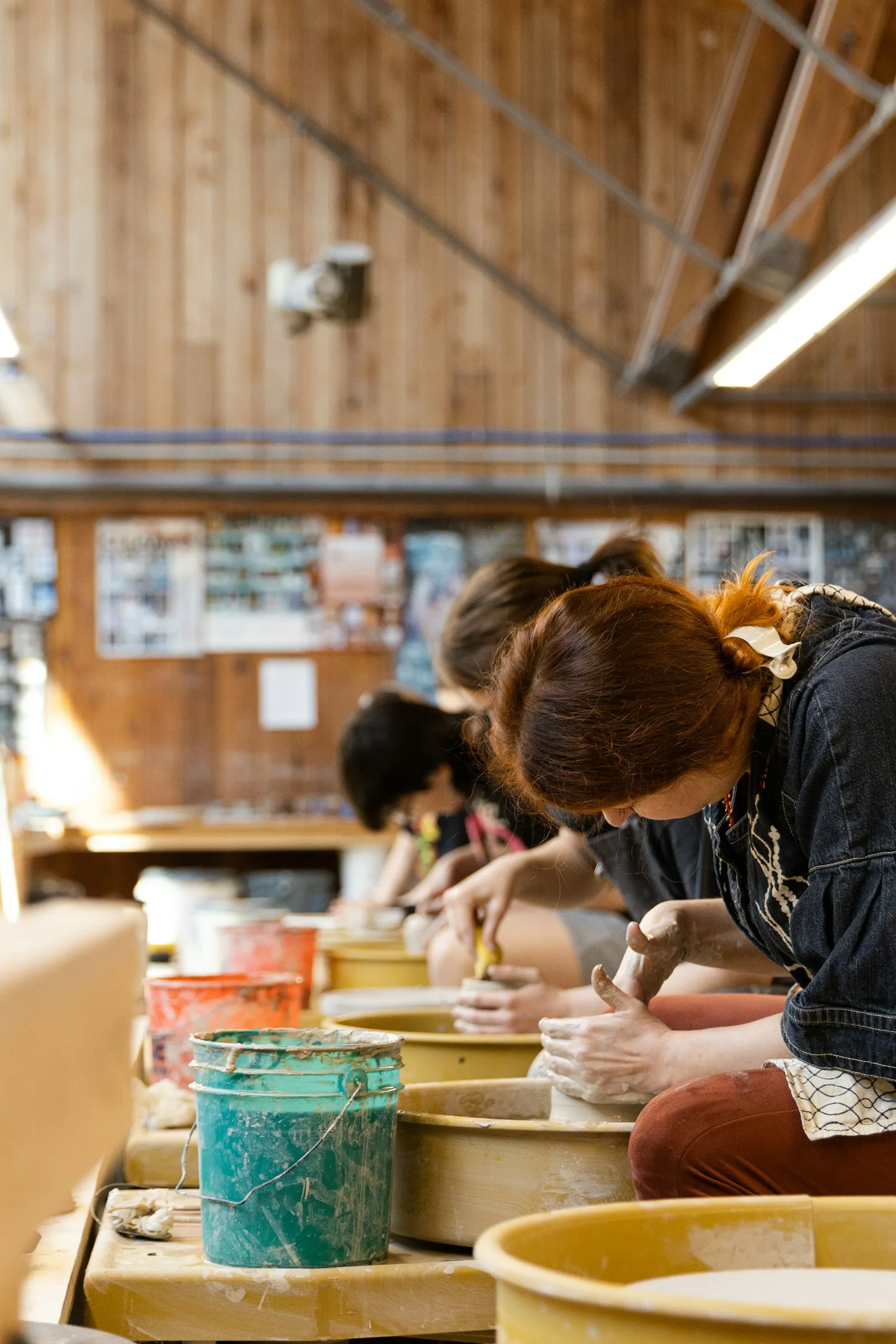 Mehrere Frauen sitzen nebeneinander an Töpferscheiben in einer hellen Werkstatt mit Holzdecke. Vorn formt eine Frau mit kupferrotem Haar konzentriert Ton, daneben stehen bunte Eimer mit Wasser.