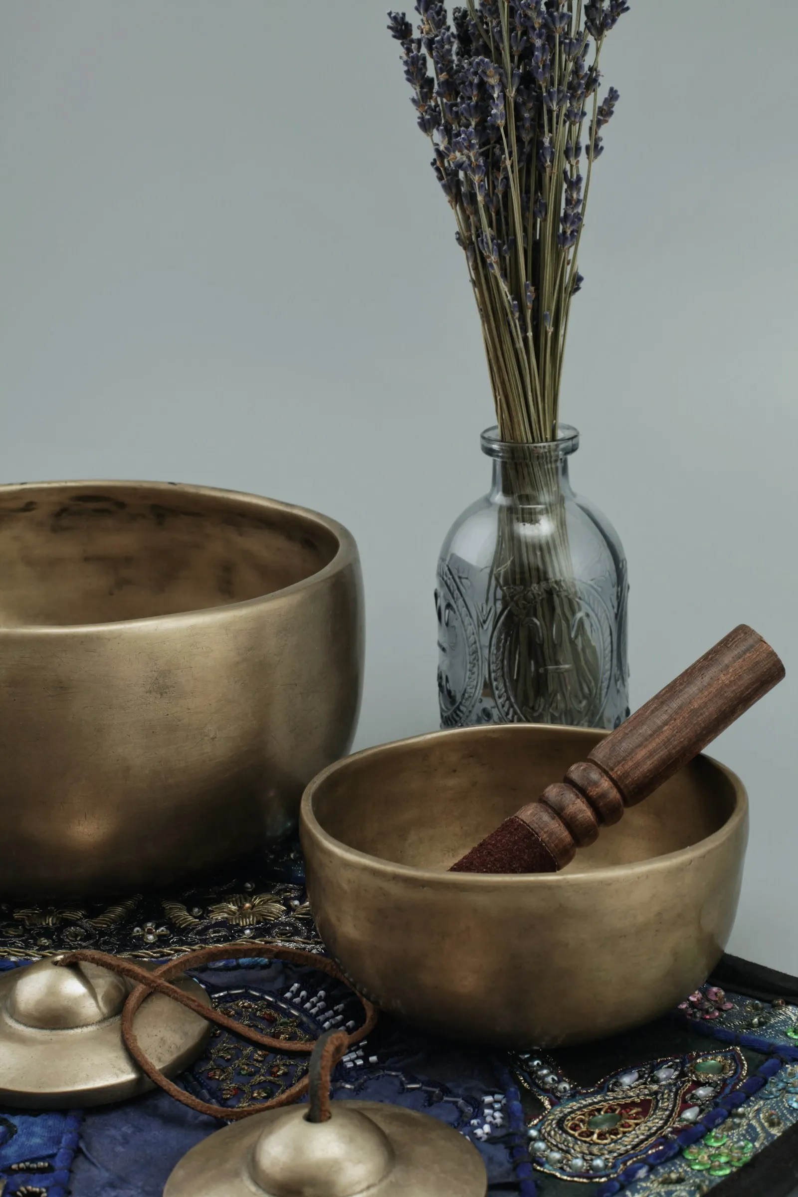 Mehrere Klangschalen mit Klöppeln, getrockneter Lavendel in einem Glasgefäß und dekorative Steine auf neutralem Untergrund