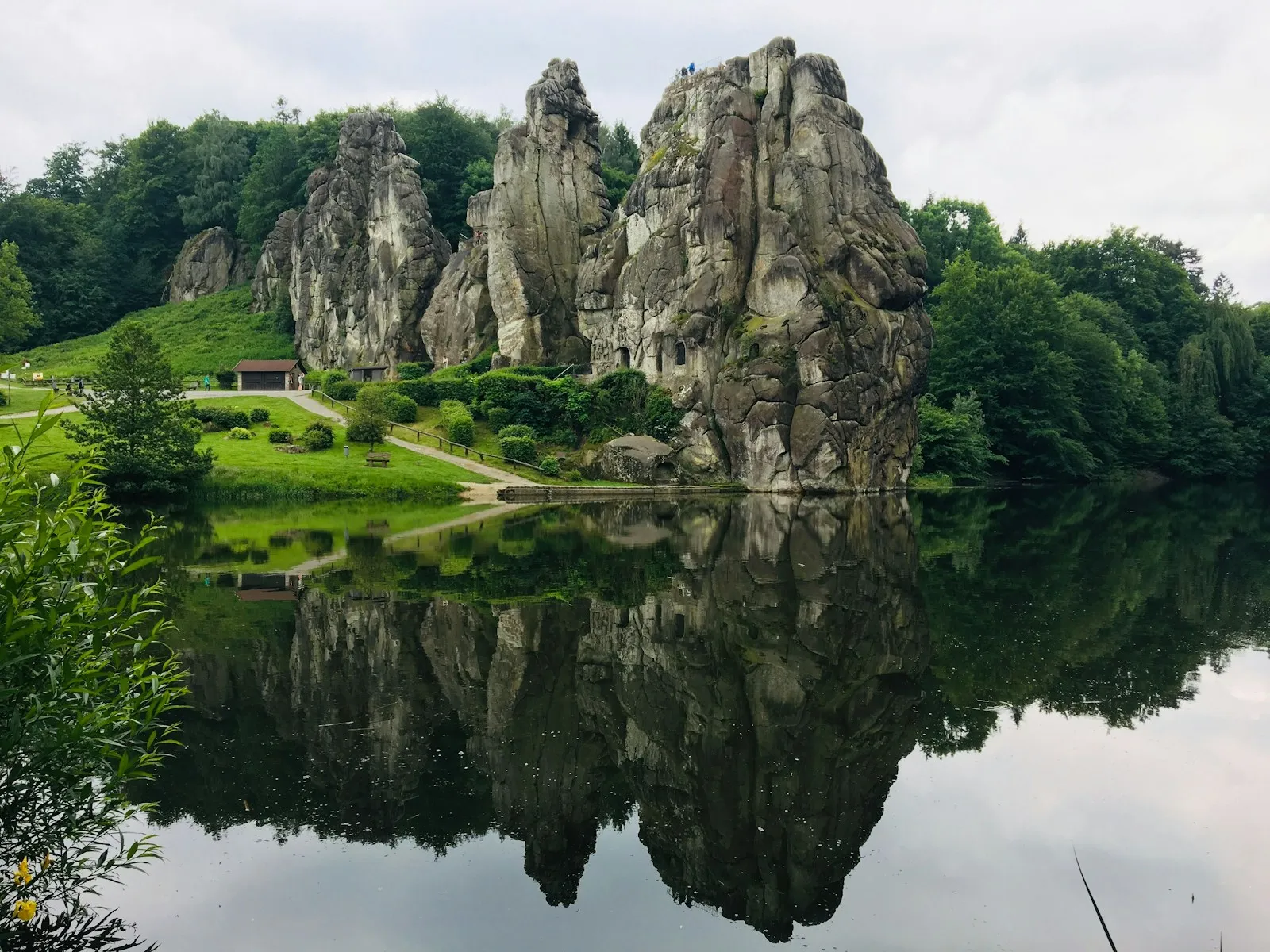 Die zerklüfteten Sandsteinfelsen der Externsteine spiegeln sich im stillen Wiembecketeich, auf dem höchsten Felskopf stehen winzig kleine Besucher am Geländer, ringsum Wiesen und Laubwald.