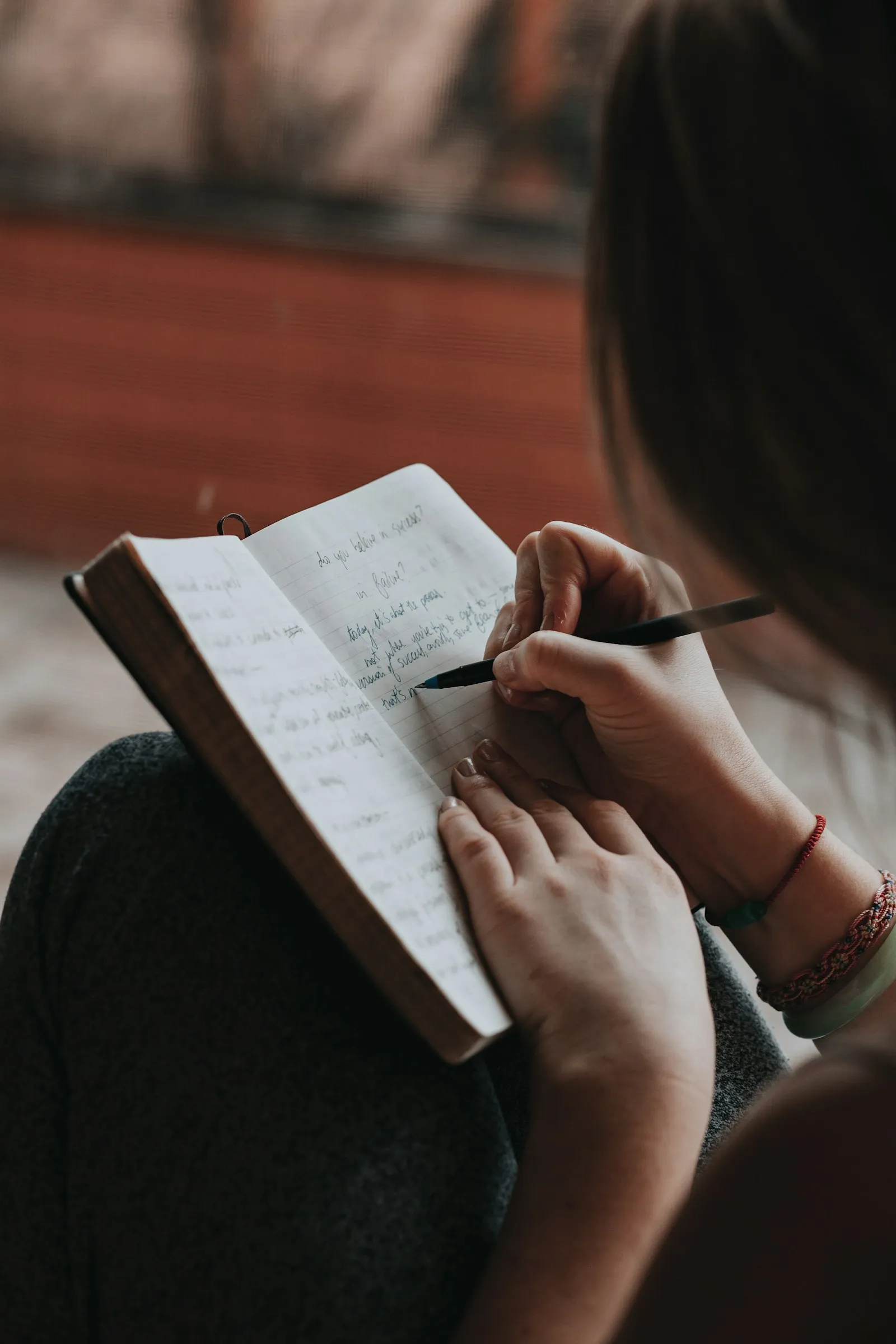 Eine Frau schreibt mit einem schwarzen Stift in ein Notizbuch, das sie in der Hand hält. Die Seiten sind mit lockerer Handschrift gefüllt, an ihrem Handgelenk trägt sie dünne Armbänder. Ihr Gesicht ist nicht zu sehen.