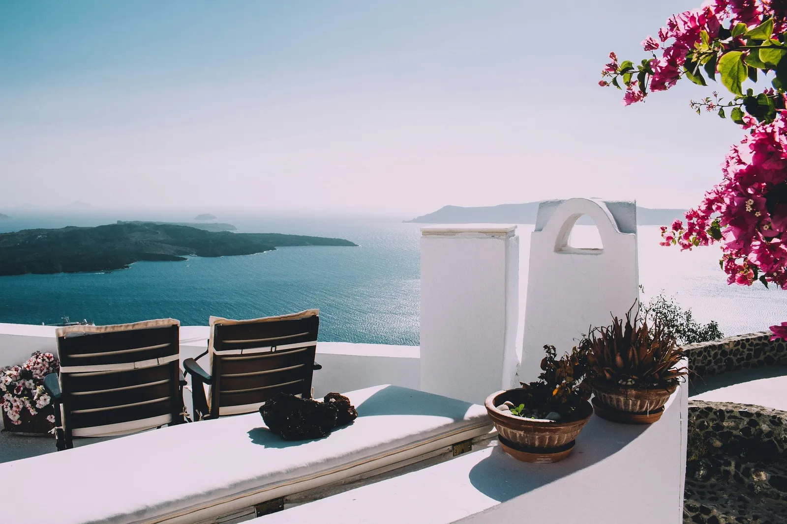 Whitewashed Santorini terrace with two wooden lounge chairs, potted plants, and bright pink bougainvillea overlooking the Aegean Sea and caldera. Luxe Mediterranean vibe.