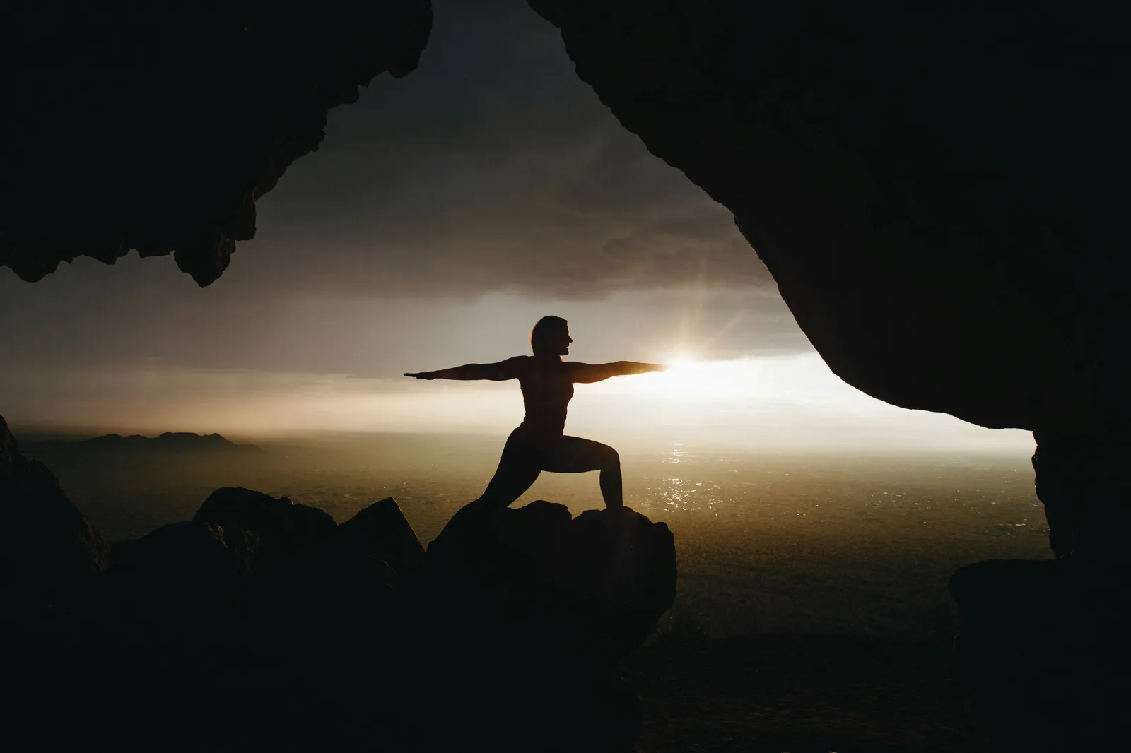 Silhouette einer Frau in der Krieger-Pose auf einem Felsvorsprung, eingerahmt von einer Höhlenöffnung im Sonnenuntergang, Berge im Hintergrund.