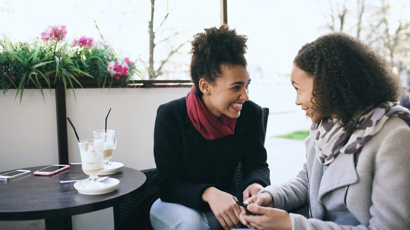 Zwei Frauen sitzen sich an einem Café-Tisch im Freien gegenüber, im Gespräch, blühende Pflanzen und Getränke auf dem Tisch.