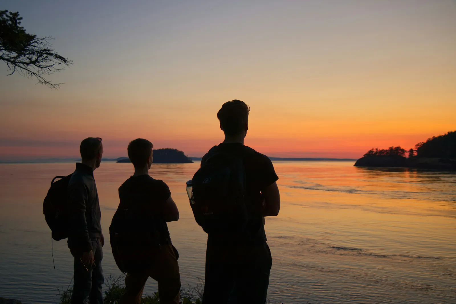 Drei Männer in Silhouette beim Sonnenuntergang über einer stillen Bucht, Rucksäcke dabei, Ende des Wandertages.