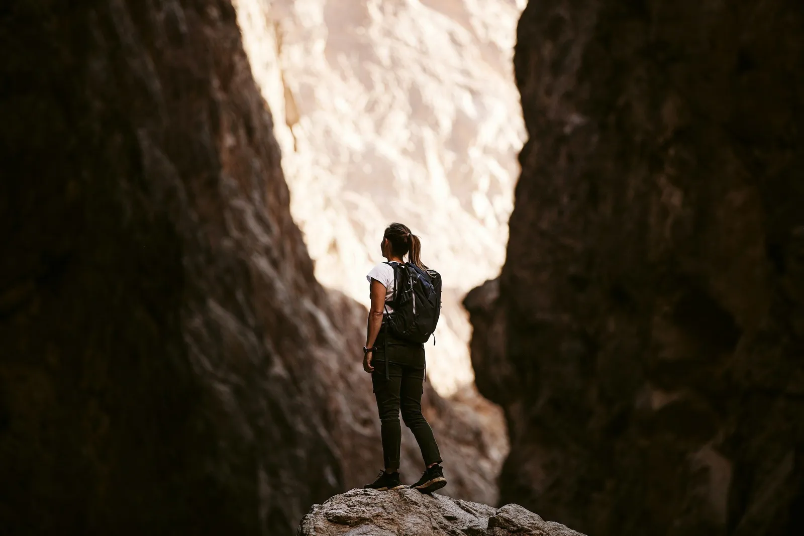 Frau mit Rucksack steht auf einem Felsen in einer engen Felsschlucht und blickt nach oben zu sonnenbeschienenen Felswänden.