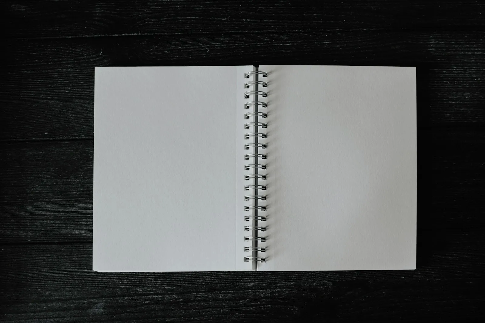 Ein aufgeschlagenes Notizbuch mit leeren weißen Seiten liegt auf einer dunklen Holzoberfläche, fotografiert von oben.