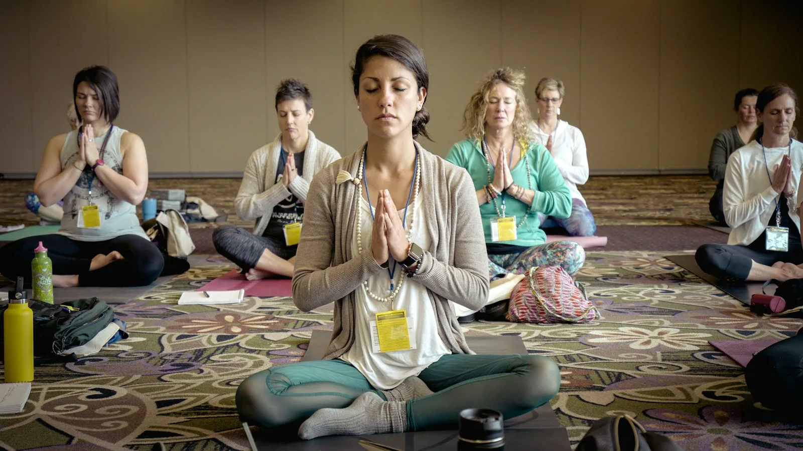 Eine Gruppe Frauen sitzt im Kreis im Schneidersitz auf Yogamatten in einem hellen Konferenzraum, Hände in Gebetshaltung vor der Brust, Augen geschlossen.