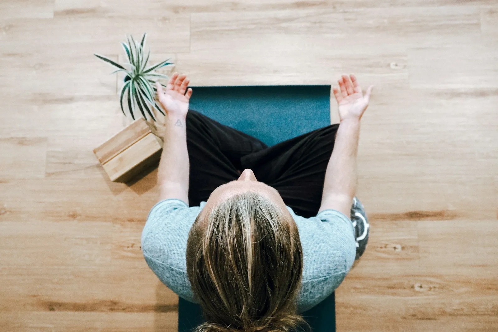 Draufsicht auf eine Frau im Schneidersitz auf einer Yogamatte auf Holzboden, daneben eine Topfpflanze und ein kleiner Bücherstapel.