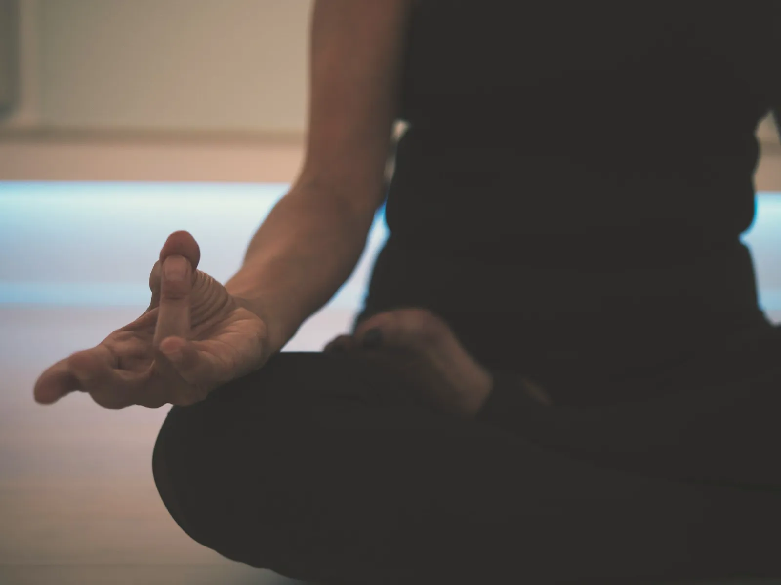 Nahaufnahme einer Person im Schneidersitz mit einer Hand in Meditations-Mudra, beleuchtet von sanftem, kühlem Licht in einem abgedunkelten Raum.