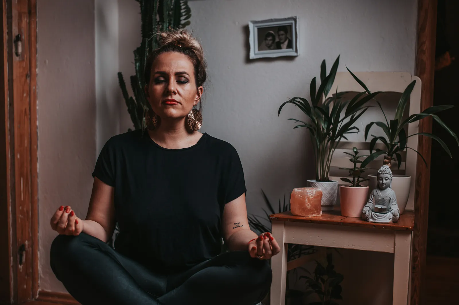Eine Frau sitzt im Schneidersitz vor einem kleinen Altar mit Buddha-Figur und Pflanzen, Augen geschlossen in konzentrierter Meditation, warmes gedämpftes Licht.