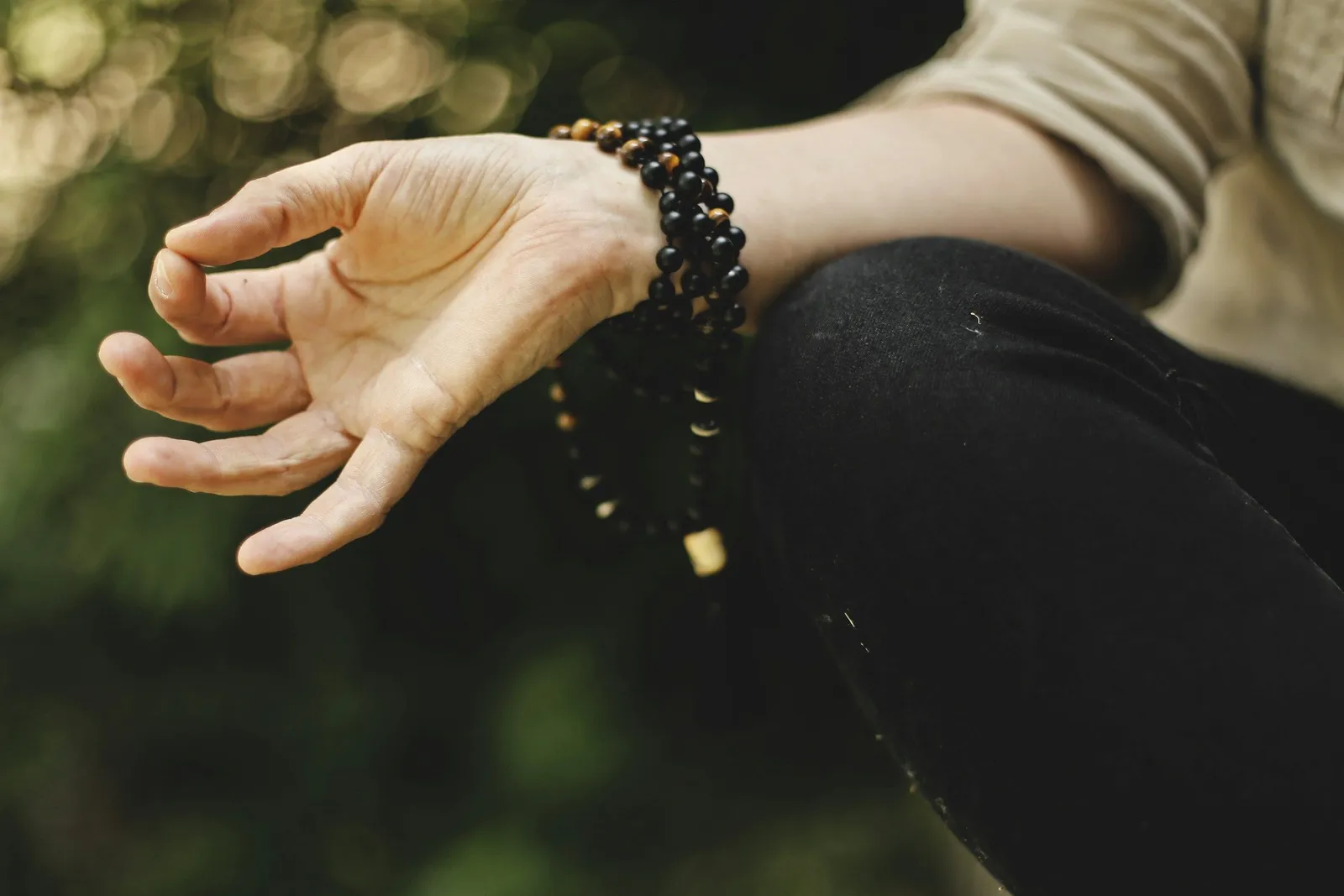 Nahaufnahme einer geöffneten Hand in Meditations-Mudra auf dem Knie, mit einer Mala-Kette aus Holzperlen, unscharfer grüner Hintergrund.
