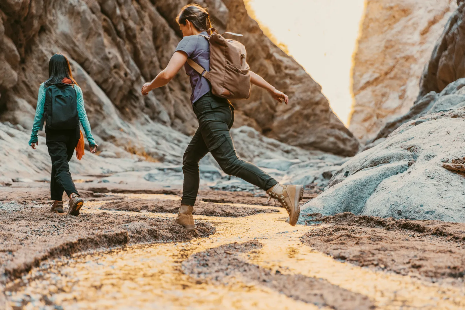 Zwei Frauen mit Rucksäcken springen lachend über Felsen in einer sonnendurchfluteten Schlucht.