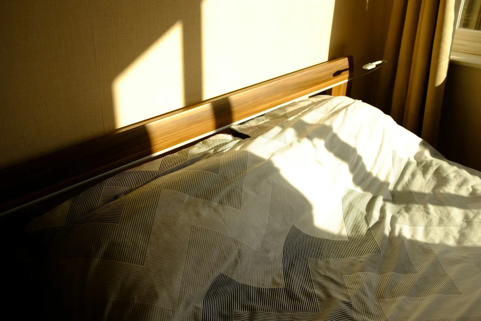 Ein zerwühltes Bett in hellem Licht, das durch Lamellenjalousien fällt und scharfe geometrische Schatten auf die Bettwäsche wirft.