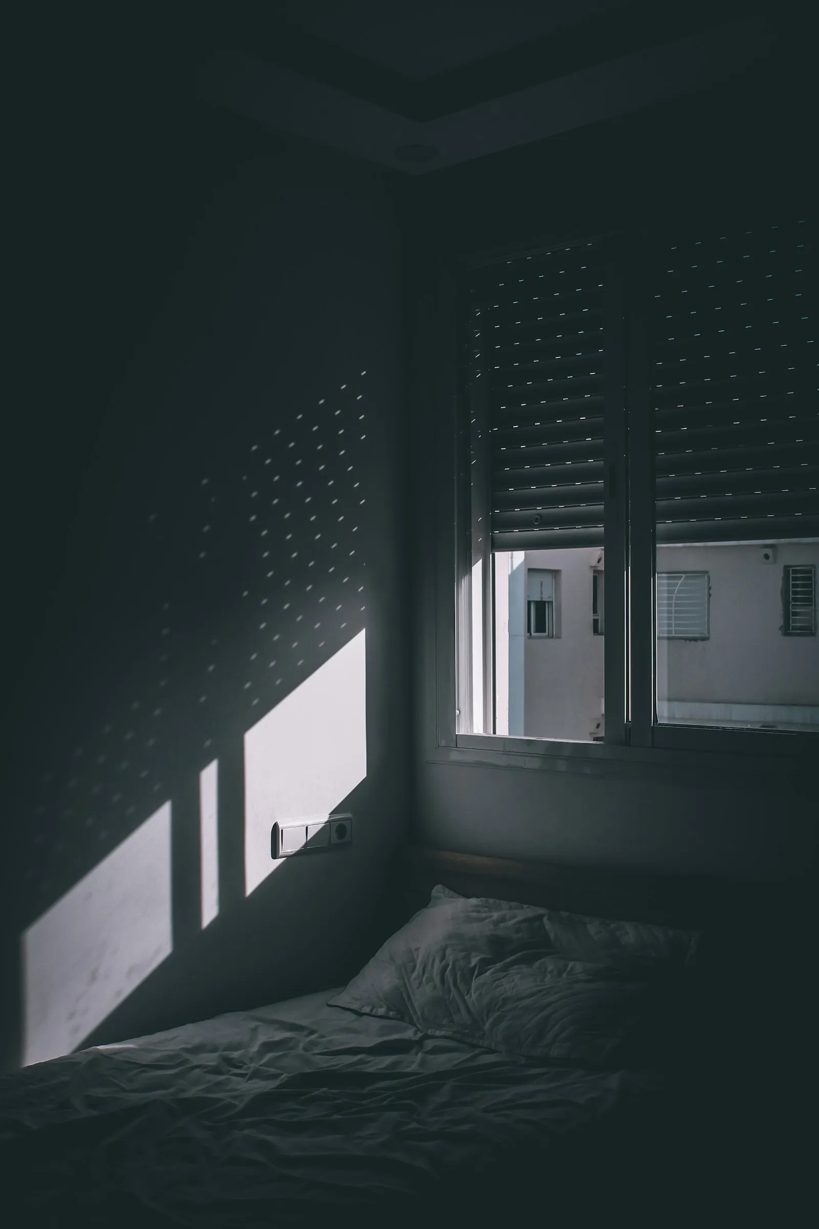 Ein dunkel bezogenes Bett im Halbdunkel, scharfe geometrische Schatten fallen durch die Jalousie auf die Bettwäsche, ein Nachbargebäude ist durchs Fenster zu erkennen.