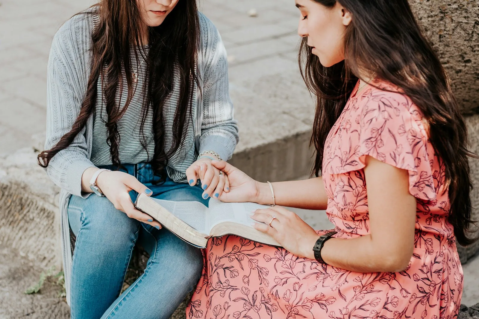 Zwei junge Frauen sitzen zusammen auf Steinstufen und lesen gemeinsam in einem aufgeschlagenen Buch, eine in einem geblümten rosafarbenen Kleid, die andere in Jeans und grauer Strickjacke. Ein stiller, konzentrierter Moment im Freien.