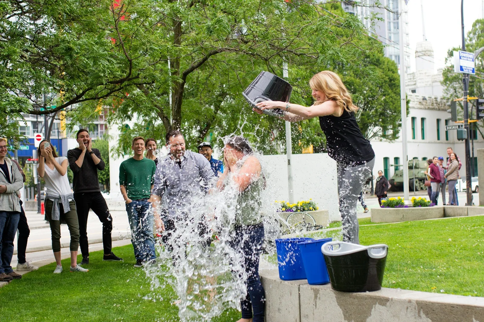 A woman pours a bucket of ice water onto a man in a suit during an ice bucket challenge, surrounded by laughing onlookers in a city plaza. Fun group team-building moment.