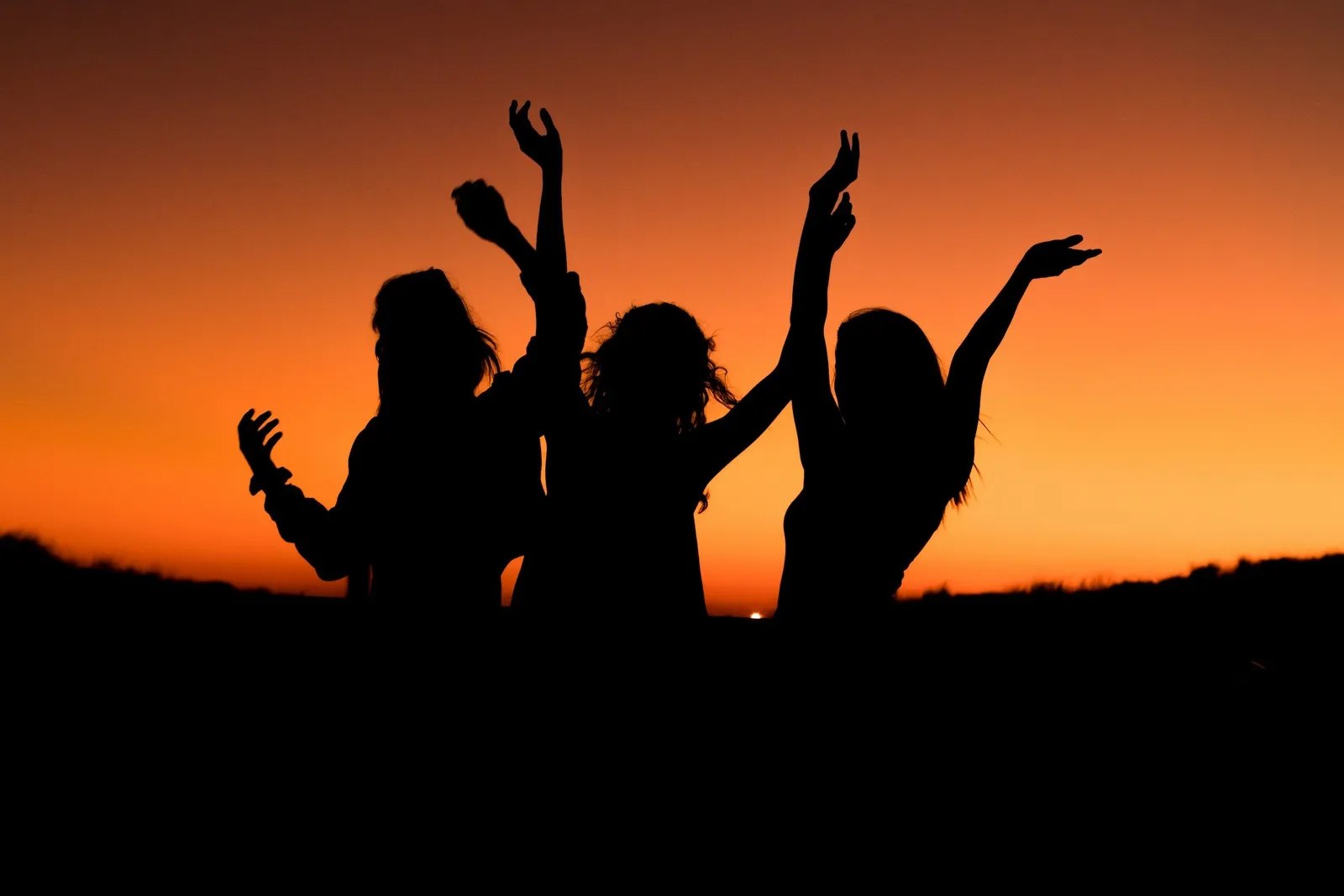 Drei Frauen als Silhouetten tanzen mit erhobenen Armen vor einem leuchtend orangefarbenen Abendhimmel, Aufbruch und Freude am Ende eines Retreats