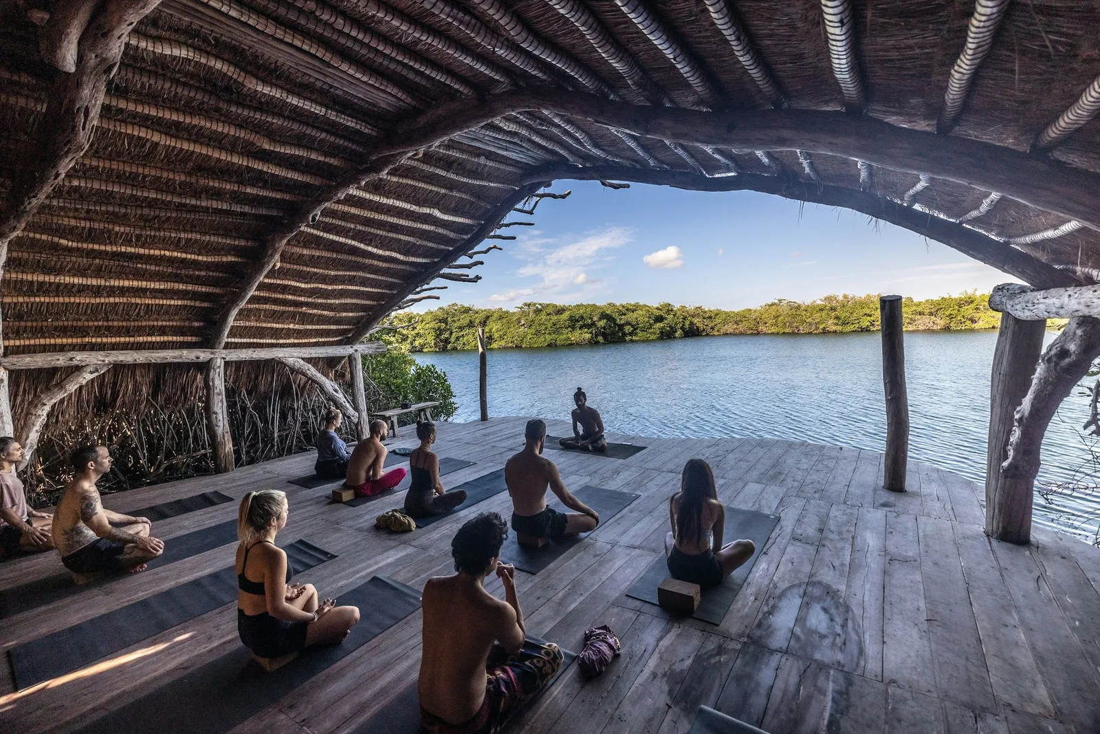Gruppe von Menschen bei der Yoga-Praxis auf einer Holzplattform unter einem Strohdach-Pavillon mit Blick auf einen ruhigen See und Mangrovenwälder.