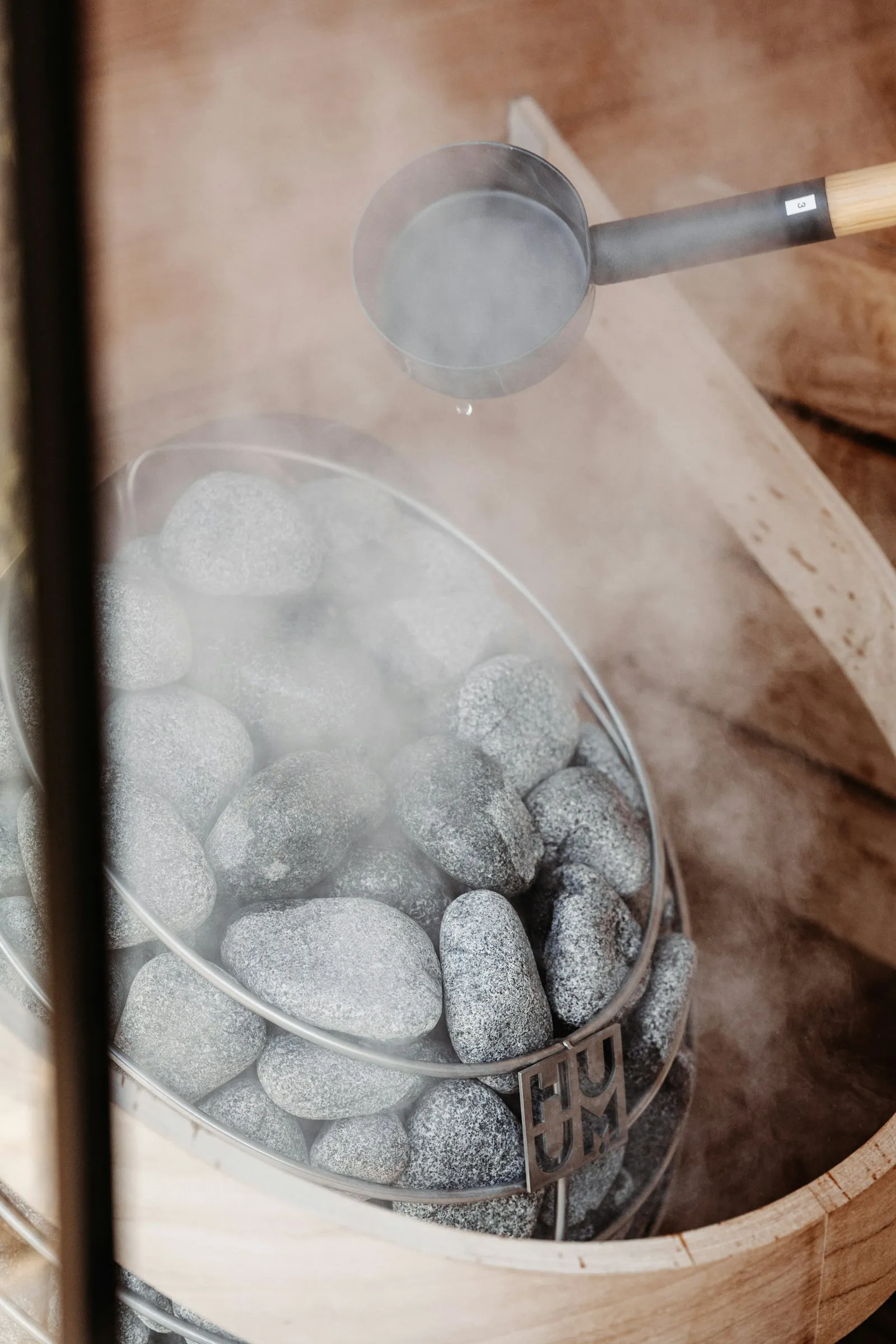 Nahaufnahme einer Metallkelle, die Wasser über heiße Saunasteine in einem Behälter gießt und dabei das zentrale Element des Sauna-Aufguss-Rituals erzeugt.
