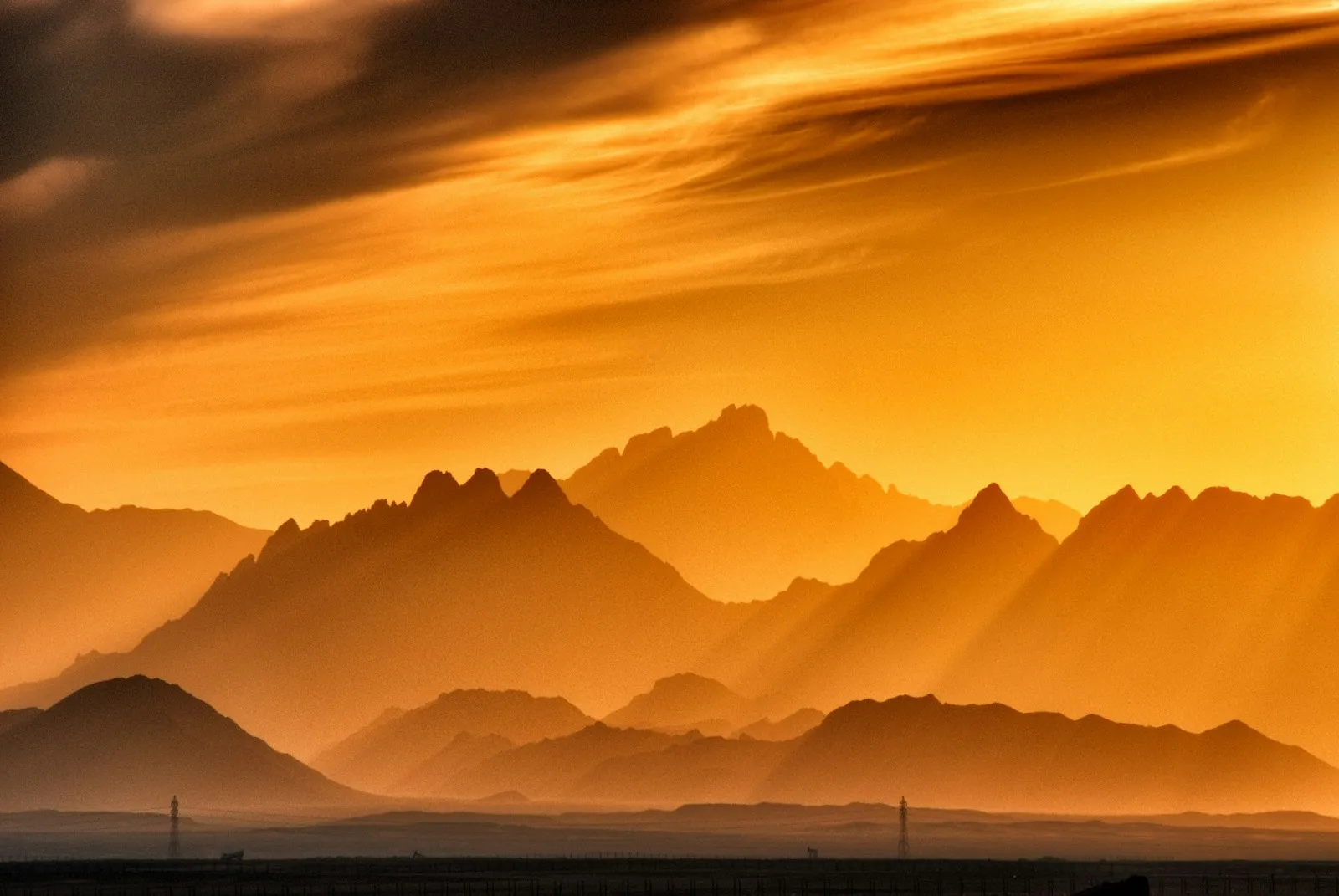 Gestaffelte Bergketten als Silhouette vor einem warmen goldenen Himmel bei Sonnenaufgang, mit atmosphärischem Dunst, der Tiefe zwischen den fernen Gipfeln schafft.